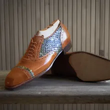 Hier sehen Sie einen Maßgefertigten Lederschuh mit braunem Glattleder und seitlichem Fischleder-Einsatz, präsentiert auf einer Werkbank.