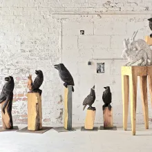 Im weissen Atelier-Raum stehen fünf Raben-Stelen und eine langbeinige Stuhl-Skulpturen, auf deren Sitzfläche ein koppolierendes Hasenpaar und auf der Lehne eine Krähe hocken.