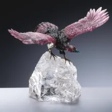 Ein Adler aus pink-grünen Turmalin mit ausgebreiteten Flügeln steht auf einem rohen, transparenten Bergkristall-Sockel.