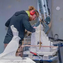 Eine Person arbeitet in einer Werkstatt an einem Steinblock. Auf dem Arbeitstisch stehen Werkzeuge und ein Messgerät zum Punktieren. Im Vordergrund ist eine kleine Steinfigur in Form eines Vogels zu sehen.