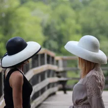 Es sind zwei Frauen in Rückenansicht auf einer Holzbrücke zu sehen. Die eine trägt ein schwarzes Kleid mit einem schwarz-cremefarbenen Strohhut, die andere ein beiges Kleid mit einem cremefarbenen Strohhut.