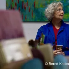 Auf dem Foto sind im Vordergrund Pinselköpfe zu sehen, im Hintergrund sitzt Ulli Zipp im blauen Malerkittel mit braunem Pappbecher in den Händen.