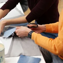 Zwei Personen sitzen an einem Tisch und bearbeiten Stoffreste. Die eine Person schneidet Jeansstoff mit einer Schere, die andere hält das Material fest.