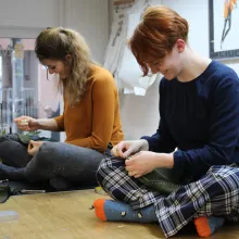 Zwei Schneiderinnen, die im Schneidersitz auf einem Tisch sitzen und nähen