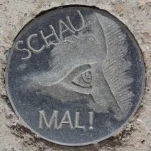 Ein Relief in dunklem Stein hier ist ein Gesichtsausschnitt- Stirn -Auge - Nase zu sehen verbunden mit dem Text - Schau mal
