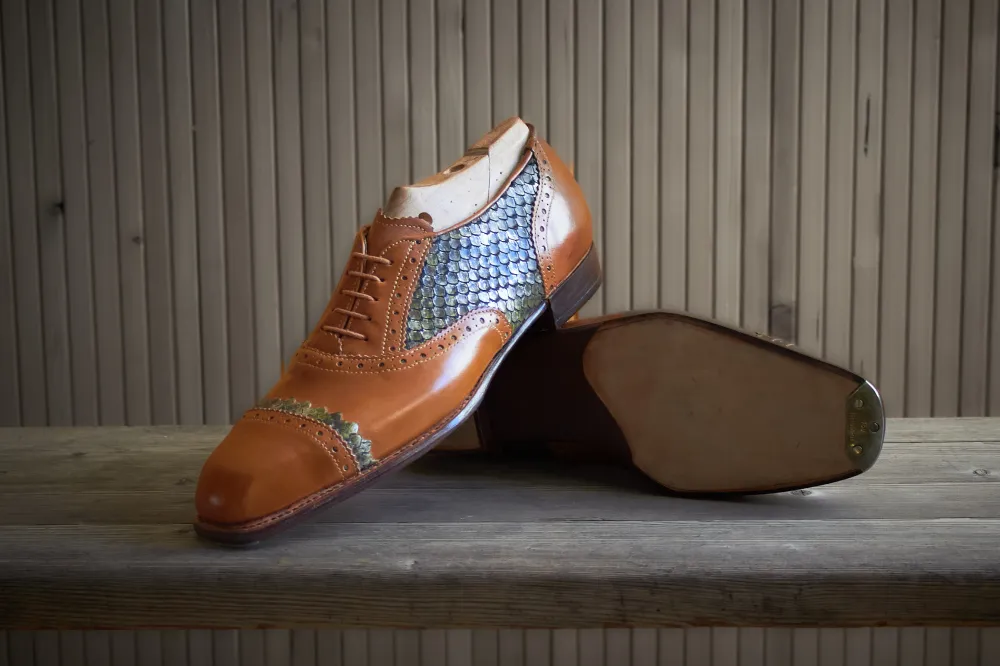 Hier sehen Sie einen Maßgefertigten Lederschuh mit braunem Glattleder und seitlichem Fischleder-Einsatz, präsentiert auf einer Werkbank.