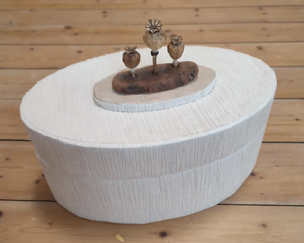 Ovale Dose aus plissiertem Papier mit Dekoration aus Rinde und Mohnkapseln