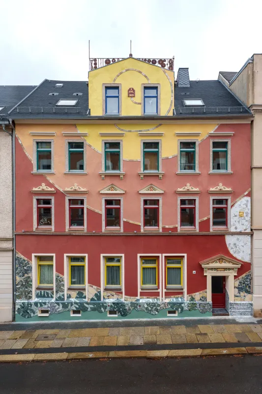 Diese Fassade wurde im letzten Jahr von mir selbst gestaltet. Es handelt sich um einen riesigen fiktiven Ammoniten, der sich in einem permanenten Farbübergang über das Haus ausbreitet. Unter dem Ammoniten wurden mit Fliesenmosaiken Monsterablätter dargestellt und eine Menge Fossilien wie Ammoniten und Fische verarbeitet. Zudem habe ich einige Fassadenteile selbst vergoldet.