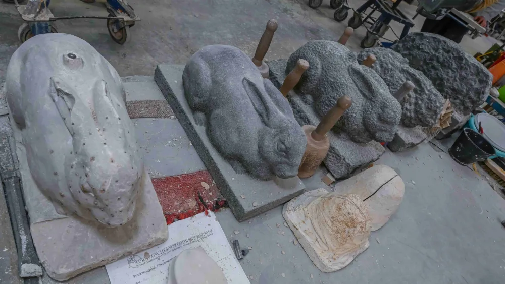 Ein Steinobjekt in Form eines Hasen ist in mehreren Arbeitsstufen zu sehen – vom grob behauenen Rohling bis zur fein ausgearbeiteten Figur. Daneben liegen Werkzeuge, die für das Modellieren und Glätten des Steins verwendet werden.