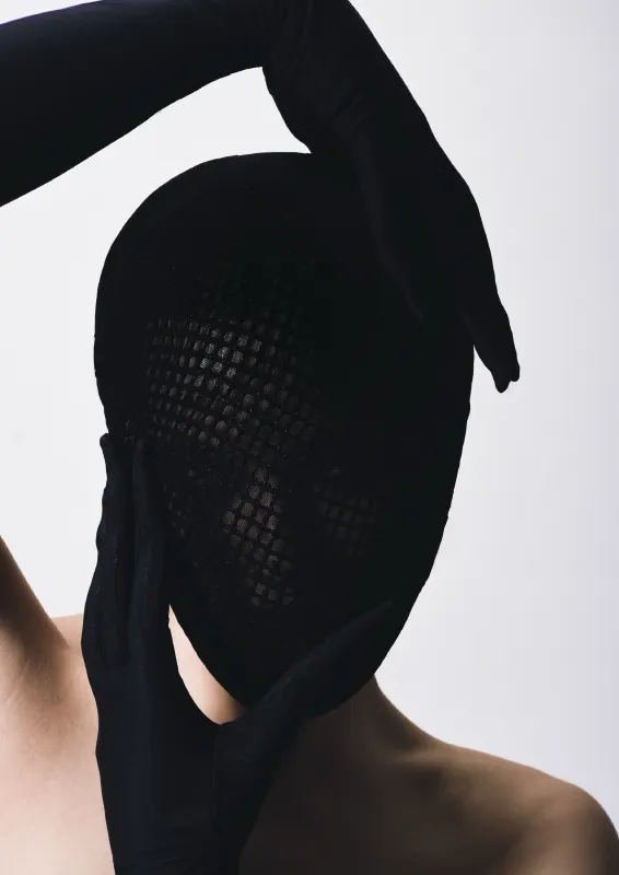 Franziska Eberle Person, die eine schwarze rasterartige Maske und schwarze lange Handschuhe trägt, die bis zum Ellenbogen reichen. Die Hände sind unten und oben am Kopf platziert.