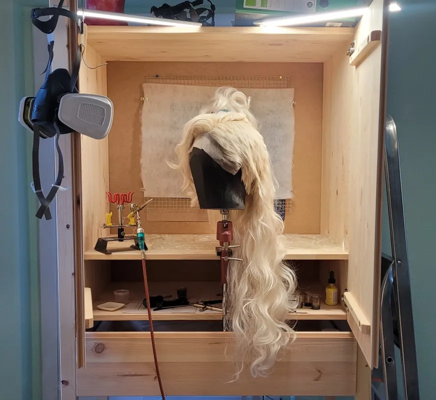 Ein beleuchteter Holzschrank mit einer langen, blonden Perücke und verschiedenen Arbeitsgeräten