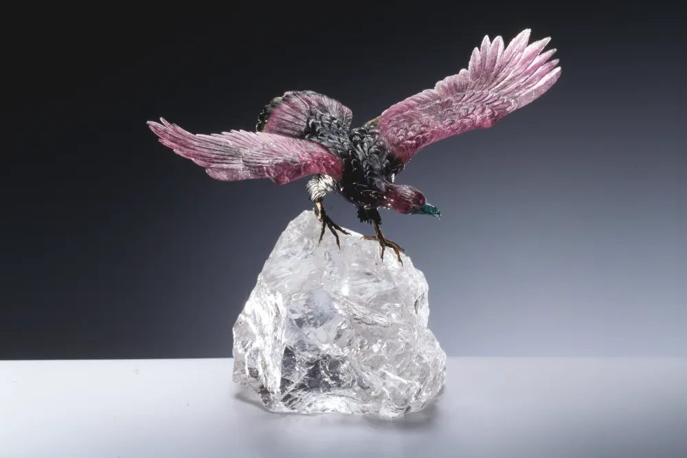 Ein Adler aus pink-grünen Turmalin mit ausgebreiteten Flügeln steht auf einem rohen, transparenten Bergkristall-Sockel.