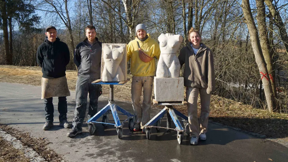 Vier Personen stehen im Freien neben zwei steinernen Torso‑Skulpturen, die auf rollbaren Arbeitsgestellen platziert sind. Die Gruppe präsentiert die fertigen oder bearbeiteten Steinwerke auf einem Weg in einer natürlichen Umgebung mit Bäumen.