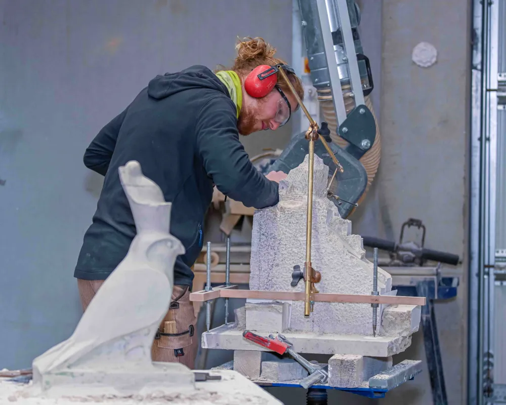 Eine Person arbeitet in einer Werkstatt an einem Steinblock. Auf dem Arbeitstisch stehen Werkzeuge und ein Messgerät zum Punktieren. Im Vordergrund ist eine kleine Steinfigur in Form eines Vogels zu sehen.