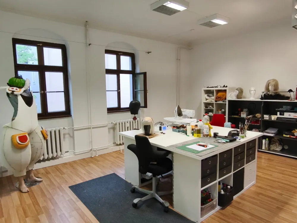 Werkstattsraum mit einem großen Mitteltisch und Regalen. In der Ecke steht ein unfertiges Kostüm auf einer Schneiderpuppe