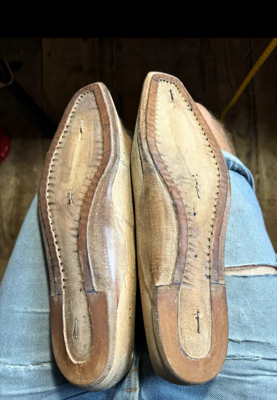 Blick auf zwei handgenähte Ledersohlen während der Fertigung, auf dem Schoß des Schuhmachers liegend.