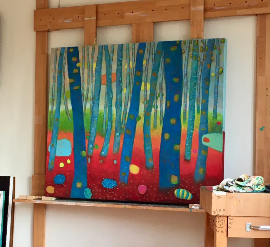 Im Atelier, auf einer Wandstaffelei steht ein großes Bild, ein Wald aus blauen Baumstämmen, der zum Hintergrund heller wird, der Waldboden ist rot, bunte Blasen schweben zwischen den Stämmen. Neben der Staffelei im Vordergrund steht ein Holztisch, darauf ein Mallappen.