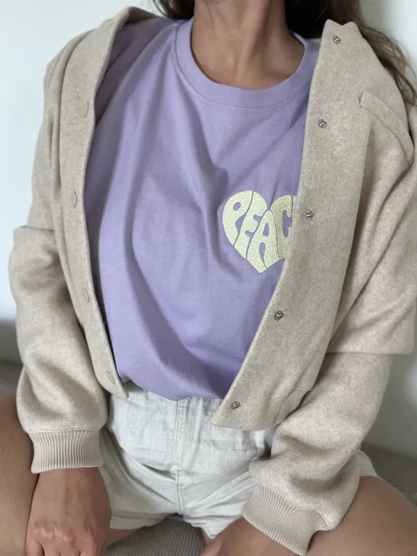 Model mit fliederfarbenem nachhaltigen T-Shirt aus Baumwolle mit einer hellgelben Stickerei der Größe 10x10 cm auf Herzhöhe, die ein Herz darstellt, das sich aus den Buchstaben PEACE ergibt