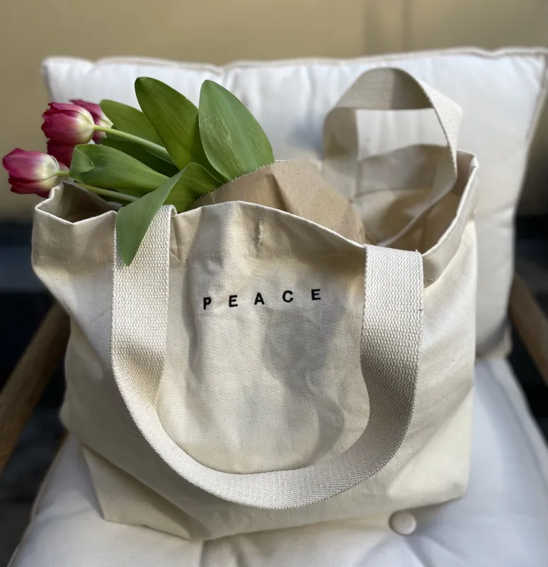 beigefarbenen Baumwoll- Canvas Tasche in Shoppergröße mit einer schwarzen minimalistischen Stickerei des Wortes PEACE in Großbuchstaben am oberen mittleren Rand der Tasche
