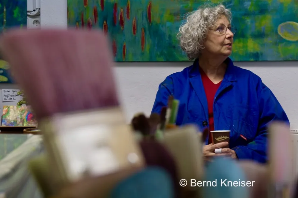 Auf dem Foto sind im Vordergrund Pinselköpfe zu sehen, im Hintergrund sitzt Ulli Zipp im blauen Malerkittel mit braunem Pappbecher in den Händen.