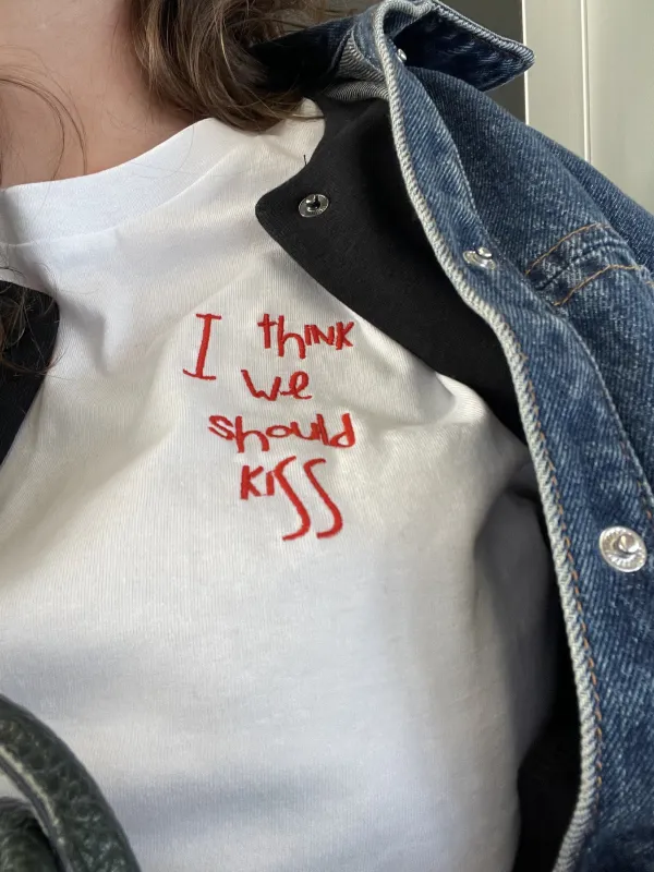 Model mit weißem T-Shirt aus Baumwolle mit einer roten Stickerei der Größe 10x10 cm auf Herzhöhe die lautet "I think we should kiss"