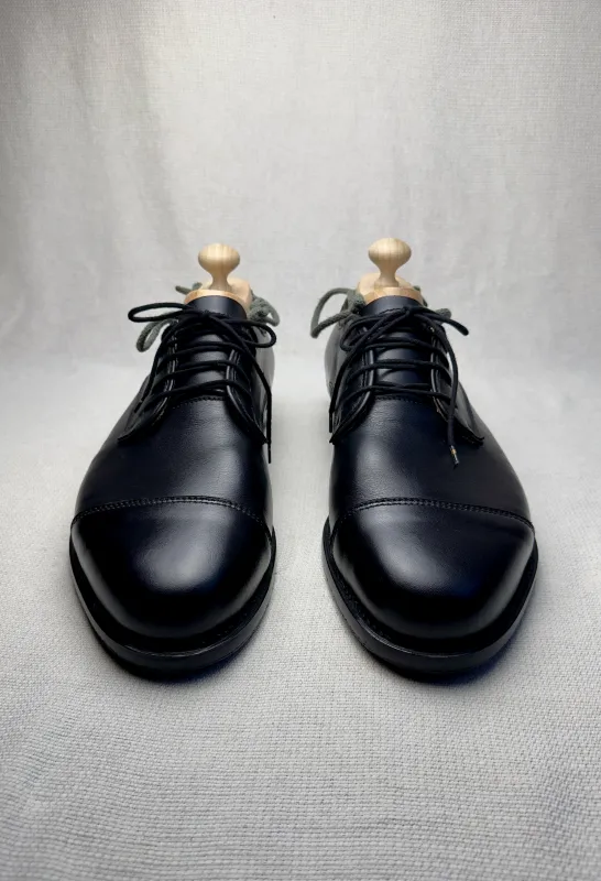 Schwarze handgefertigte Maßschuhe mit Schuhformern, frontal fotografiert auf hellem Stoffhintergrund. 