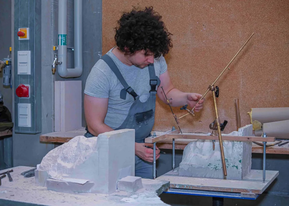 Eine Person arbeitet in einer Werkstatt an einem Steinblock. Auf dem Tisch steht ein Punktiergerät mit Metallstangen, das zur genauen Übertragung von Maßen dient. Rundherum liegen Werkzeuge und weiterer Stein, der bearbeitet wird.