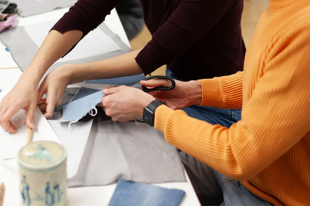 Zwei Personen sitzen an einem Tisch und bearbeiten Stoffreste. Die eine Person schneidet Jeansstoff mit einer Schere, die andere hält das Material fest.