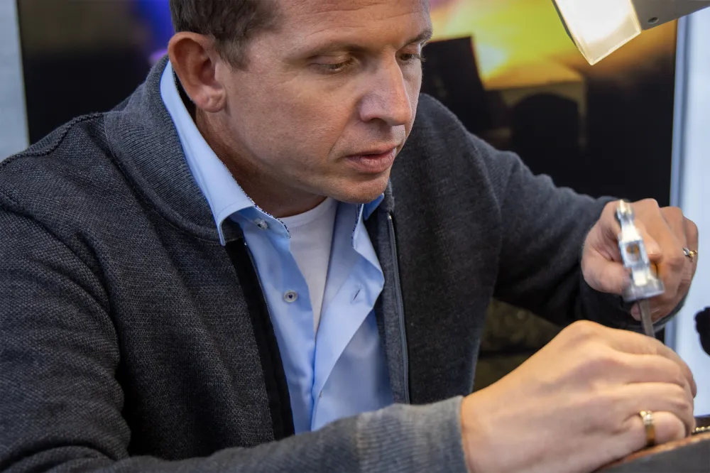 Goldschmied bei der Arbeit in seiner Werkstatt: Mit einem kleinen Hammer bearbeitet er konzentriert ein Schmuckstück im Schraubstock unter einer Lupenleuchte.