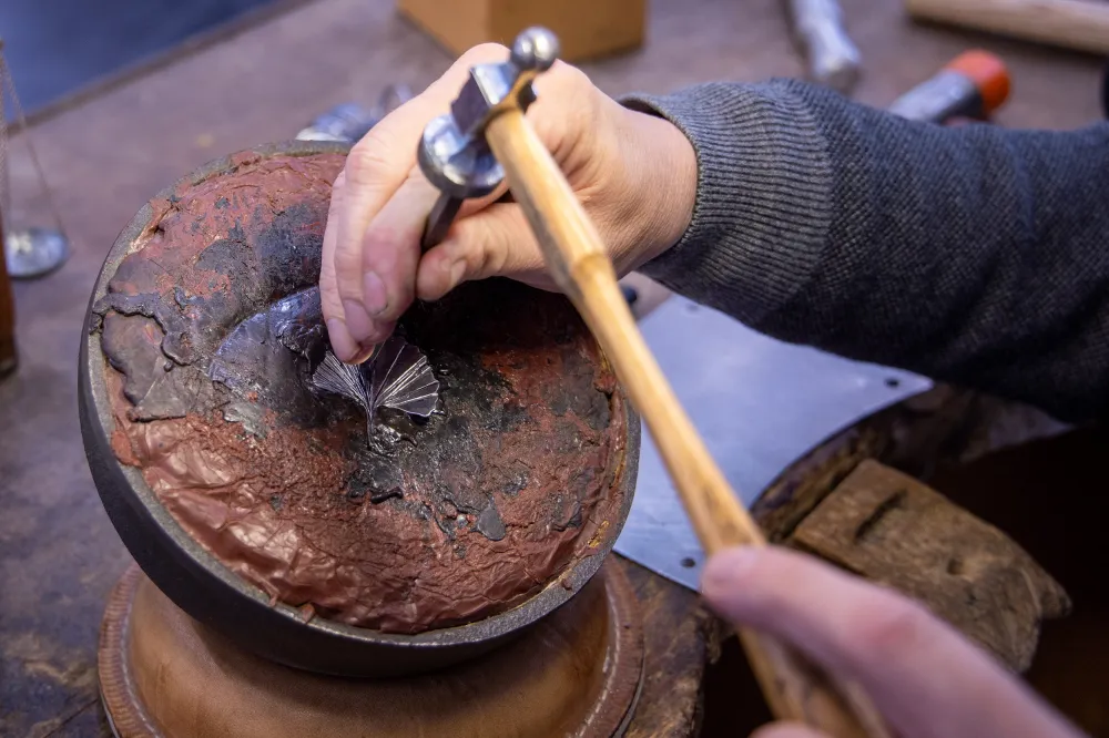 Goldschmied ziseliert ein silbernes Ginkgoblatt mit Ziselierhammer und Punze auf einer Kittkugel in der Werkstatt von Goldschmiede Mandat.