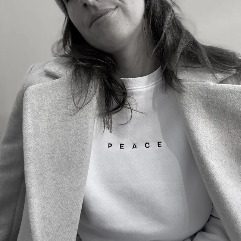 weißes Sweatshirt aus Baumwolle mit einer schwarzen minimalistischen Stickerei des Wortes PEACE in Großbuchstaben vorne, mittig, kurz über der Brust des Models