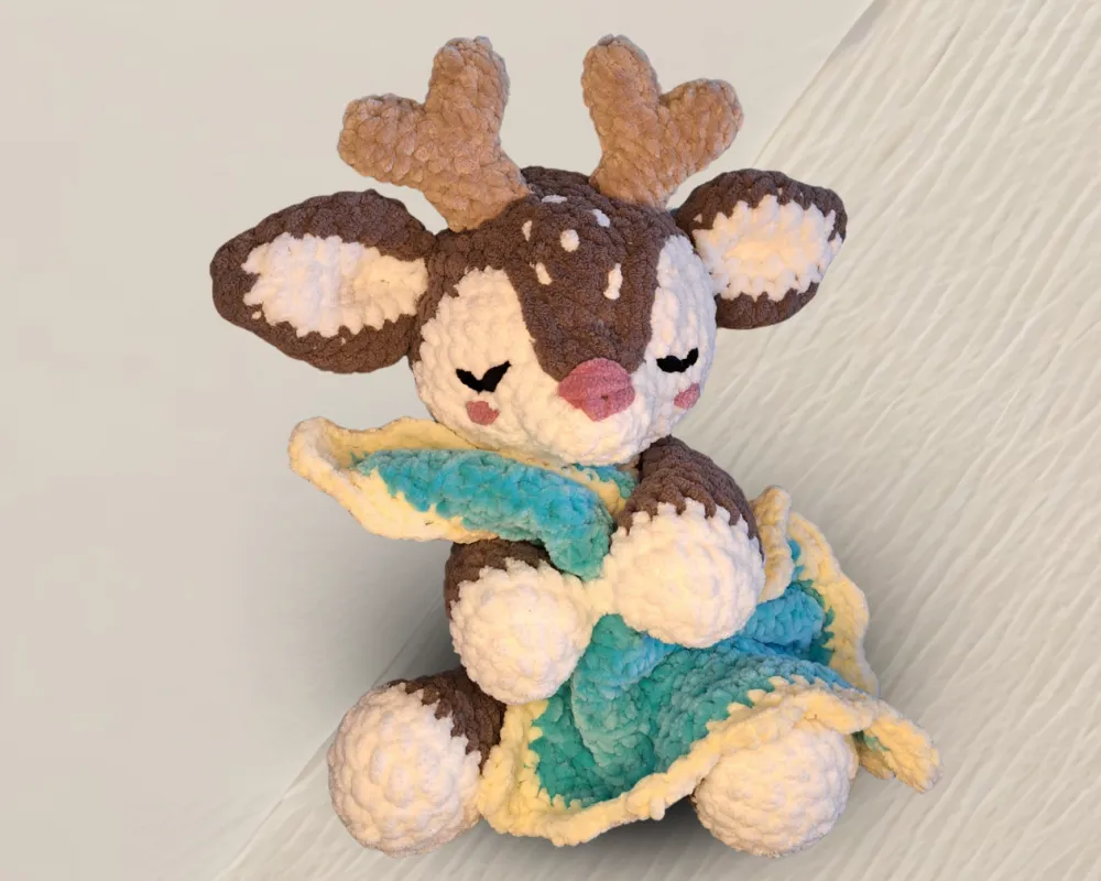 Ein gehäkeltes Kuscheltier-Reh mit süßem Geweih, dessen Augen geschlossen sind und es kuschelt eine Decke in der Farbe Türkis mit gelbem Rand