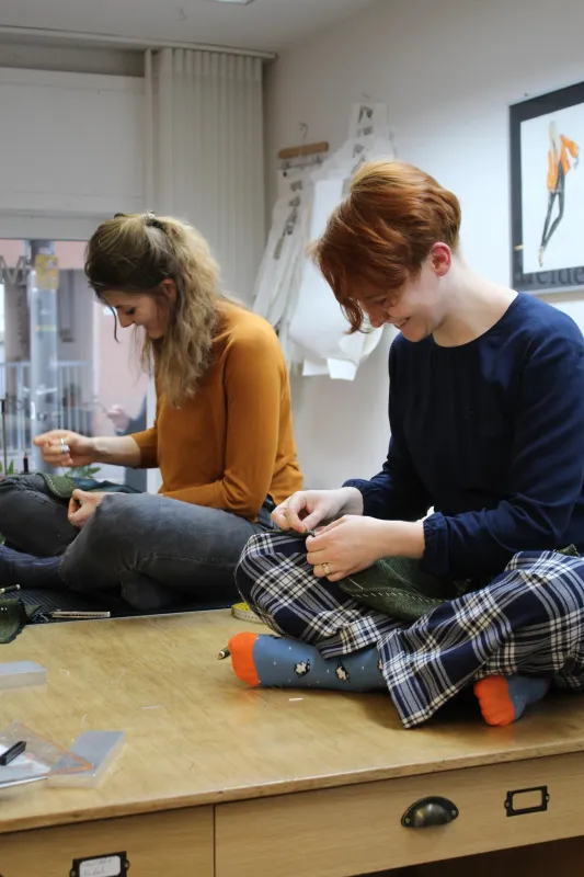 Zwei Schneiderinnen, die im Schneidersitz auf einem Tisch sitzen und nähen