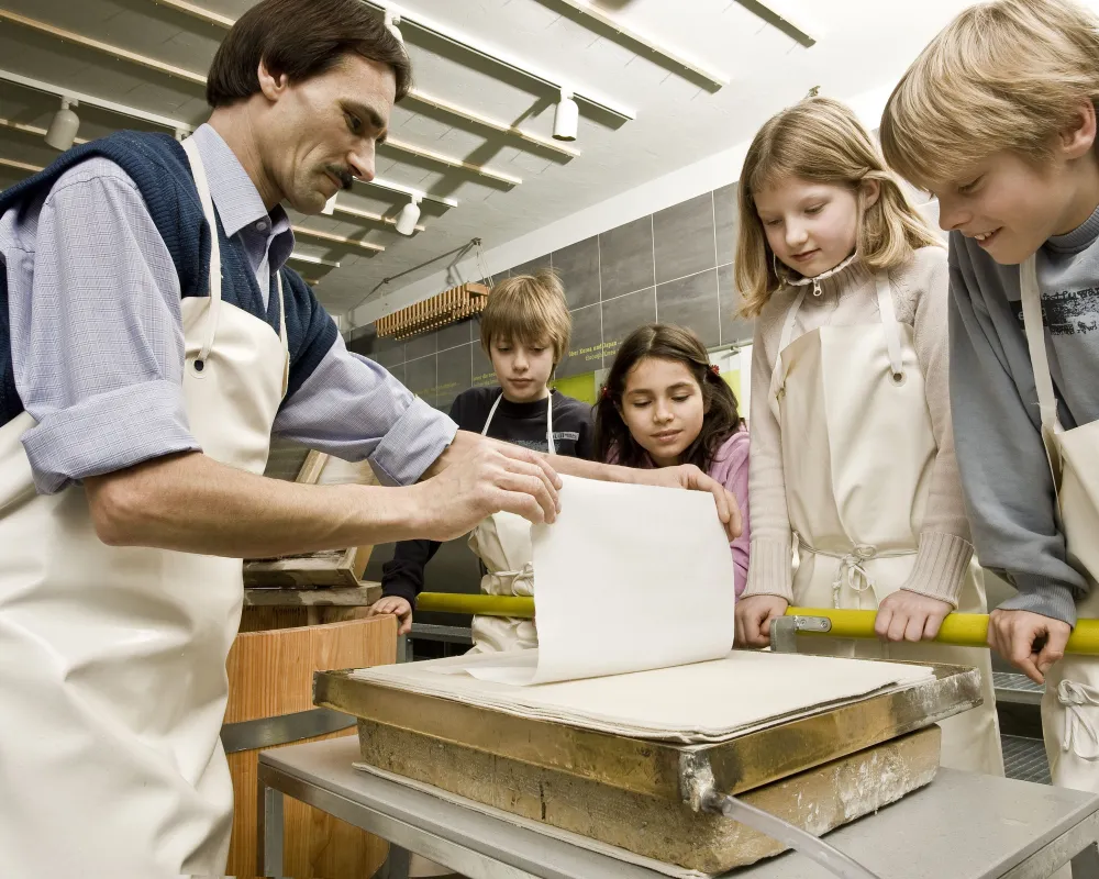 Ein Mann hält einen frisch geschöpften Bogen Papier hoch. Mehrere Kinder schauen zu.