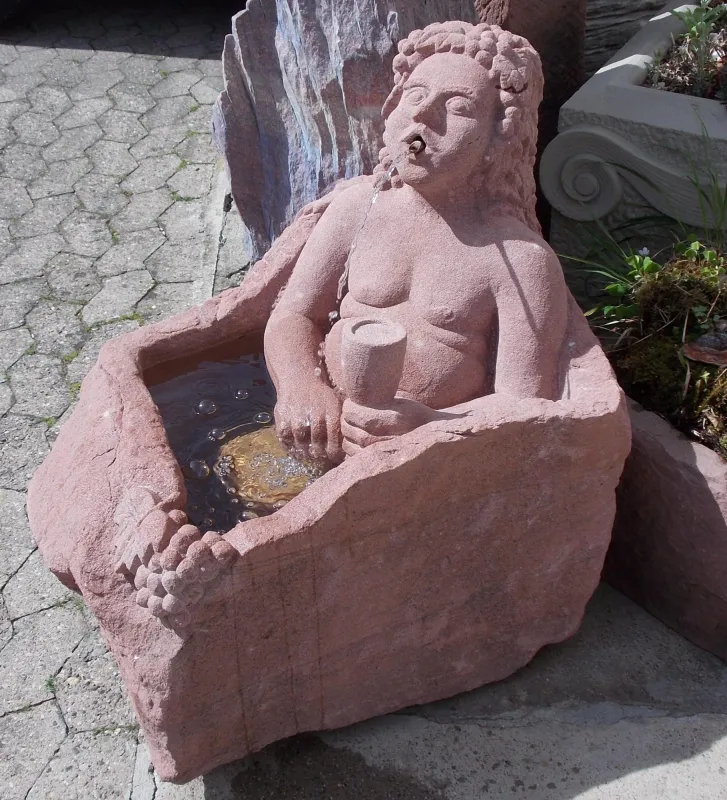 Brunnen in dem ein Mann sitzt welcher ein Becher in der Hand hält. Er spritzt Wasser aus dem Mund in den Trog