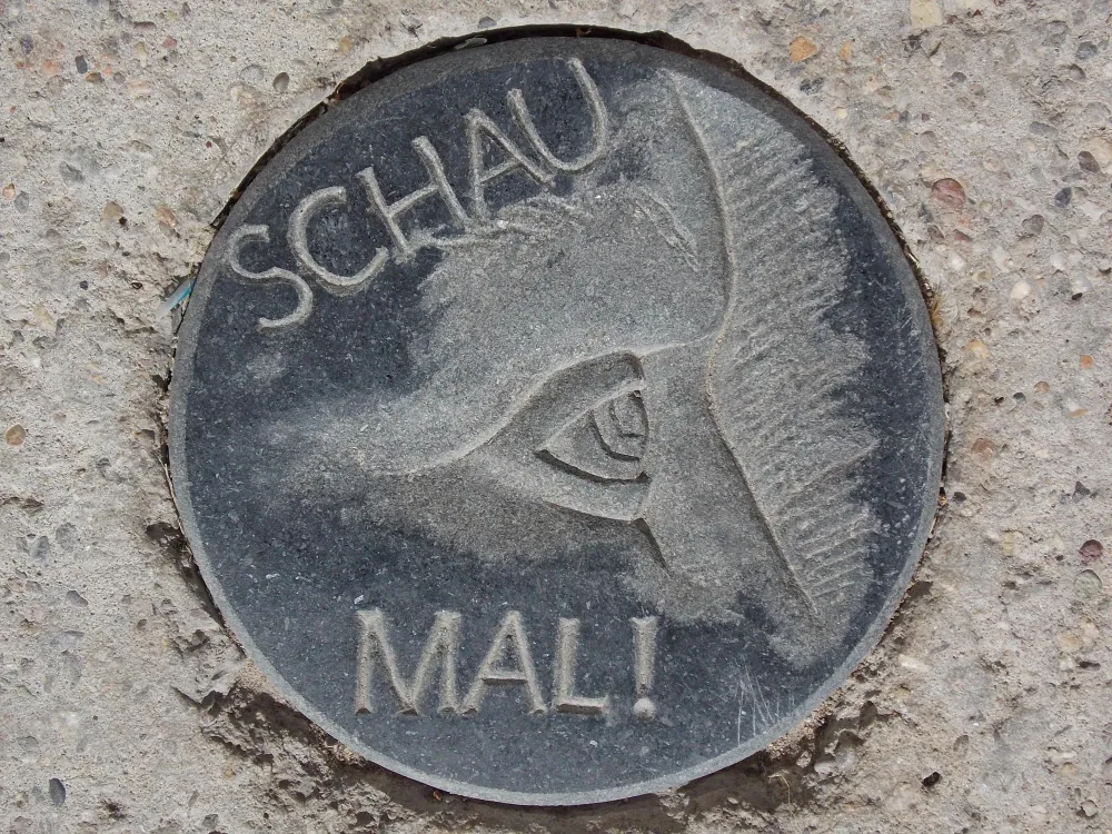 Ein Relief in dunklem Stein hier ist ein Gesichtsausschnitt- Stirn -Auge - Nase zu sehen verbunden mit dem Text - Schau mal