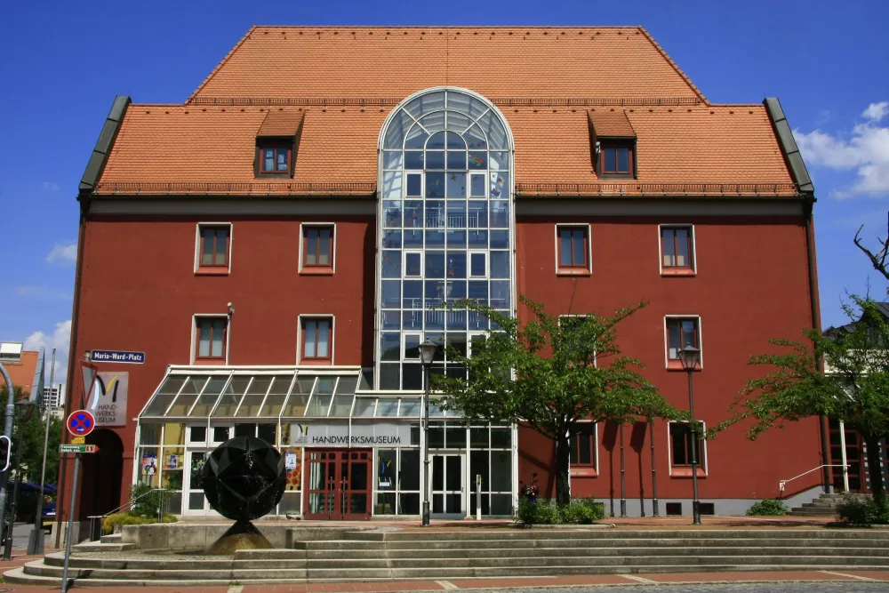 Stadt Deggendorf/Rudi Scharf Rotes Gebäude mit Glasvorbau. Auf dem Platz davor befinden sich Bäume und ein Brunnen.