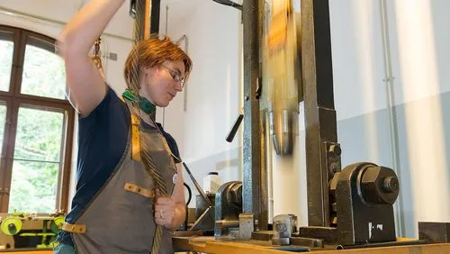 Fallhammer in der Schmuckmanufaktur Eine Frau mit Arbeitsschürze zieht an dem Seil des Fallhammers. Das Gewicht wird dadurch in der Führung nach oben gezogen, bevor es auf das Gesenk fällt.